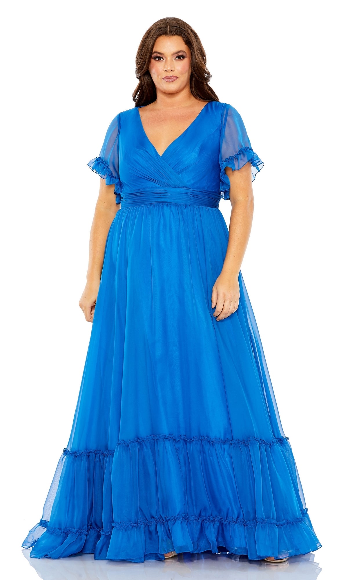 A woman stands against a white background wearing the Long Plus-Size Formal Dress 68430 by Mac Duggal, a royal blue gown with a v-neckline, short sheer sleeves, and a ruffled hem.